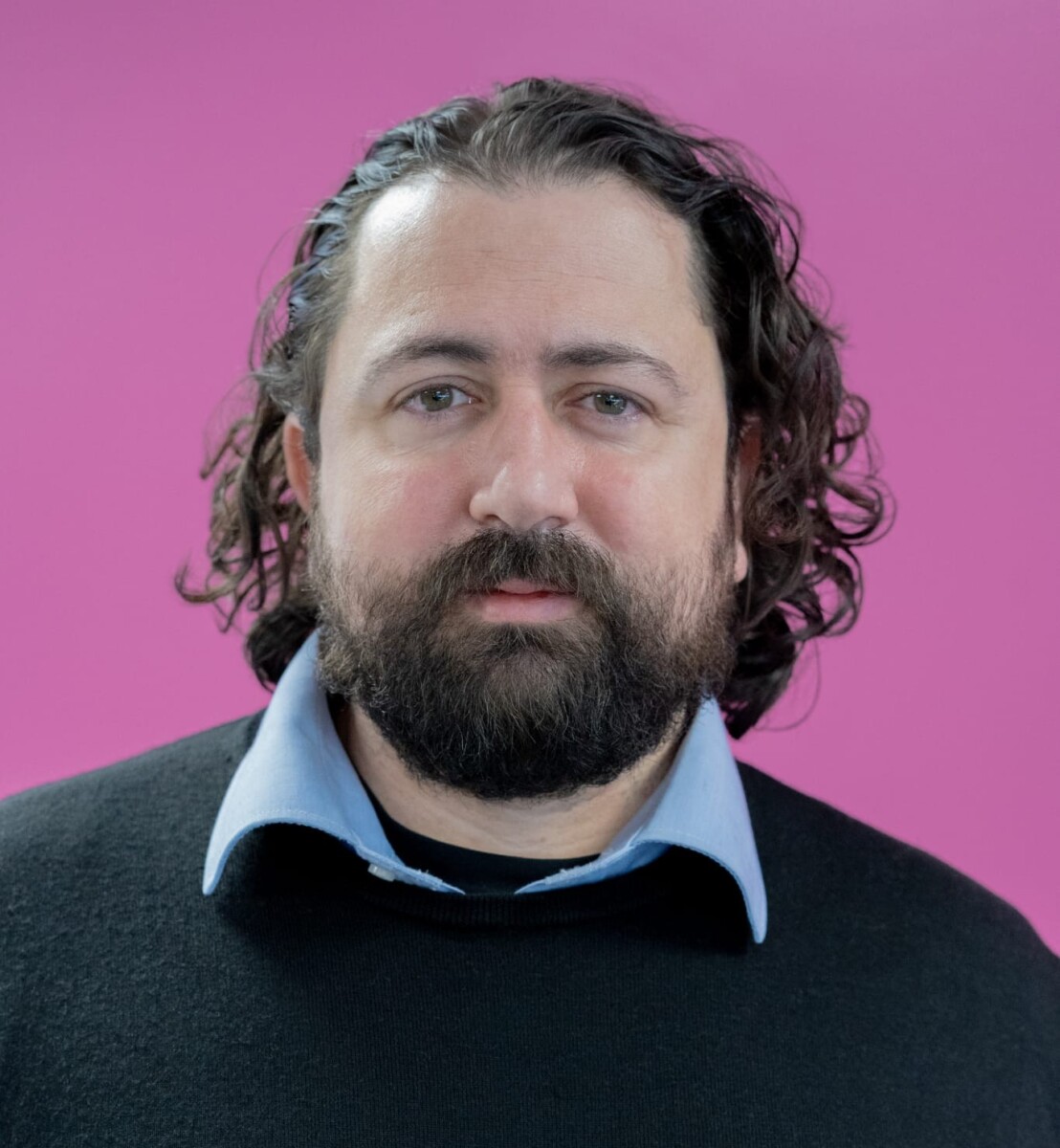 Mies, jolla on tummat kiharat hiukset ja parta ja jolla on musta villapaita vaaleansinisen kauluspaidan päällä, seisoo vaaleanpunaisen taustan edessä.