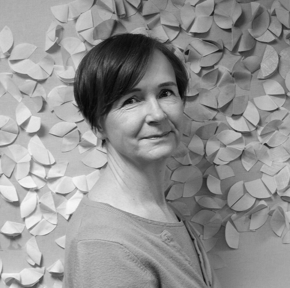 Lyhythiuksinen nainen, jolla on vaalea villapaita, hymyilee lempeästi kameralle. Hän seisoo päällekkäisillä paperilehdillä koristellun seinän edessä. Kuva on mustavalkoinen.