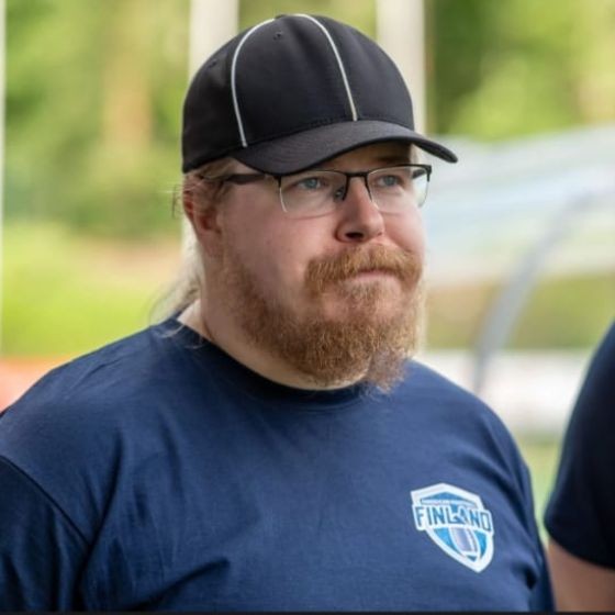 Ulkona seisoo parrakas, silmälasipäinen ja mustapäällysteinen henkilö, jolla on tummansininen t-paita, jossa on Suomen logo. Taustalla on hämärtynyt vihreä lehvistö ja rakenteet näkyvät.