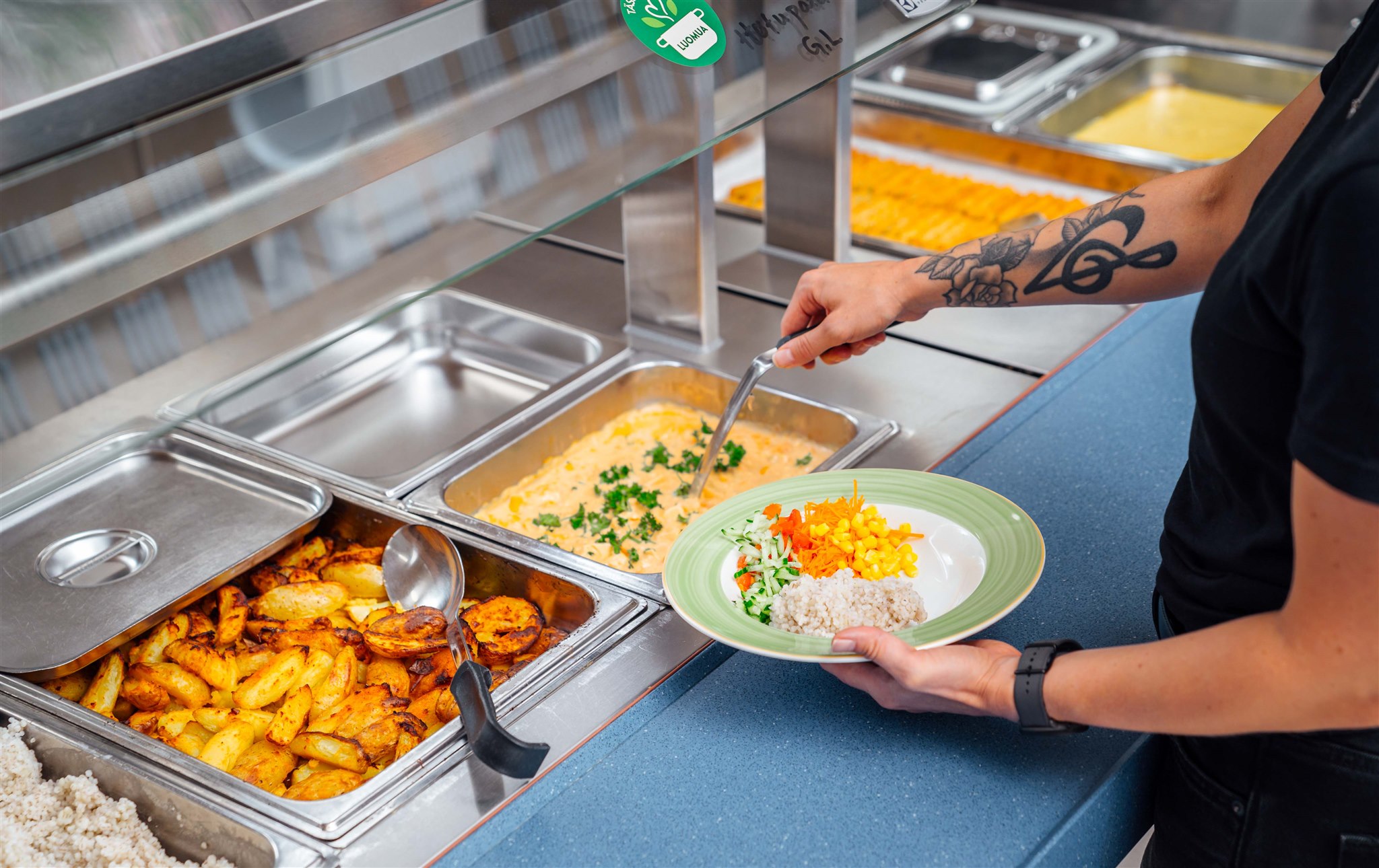Henkilö tarjoilee ruokaa kanttiinityylisestä noutopöydästä, pitää lautasella riisiä, vihanneksia ja kauhoo kermaista ruokaa seisoessaan paahdettujen perunoiden ja muiden kuumien ruokien tarjottimien vieressä.