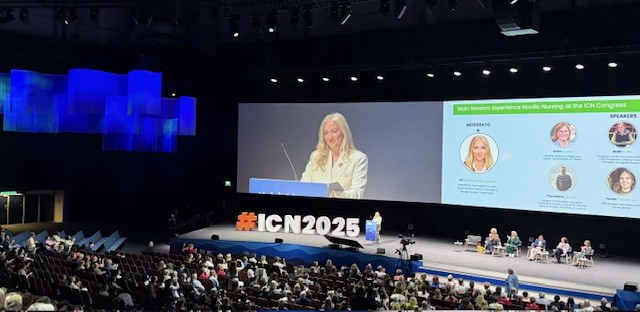 Nainen puhuu korokkeella suurella lavalla konferenssissa. Hänen takanaan olevalla näytöllä näkyy ICN2025 ja puhujien kuvia. Yleisö istuu lavaa vastapäätä tilavassa, modernissa auditoriossa.