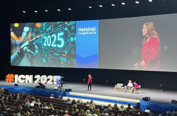 Suuri konferenssisali, lavalla puhuja, näytöllä "2025" robottikädellä ja teksti "Helsinki Congrès du CII". Yleisö istuu isojen kirjainten alla, joissa lukee "#ICN2025.".