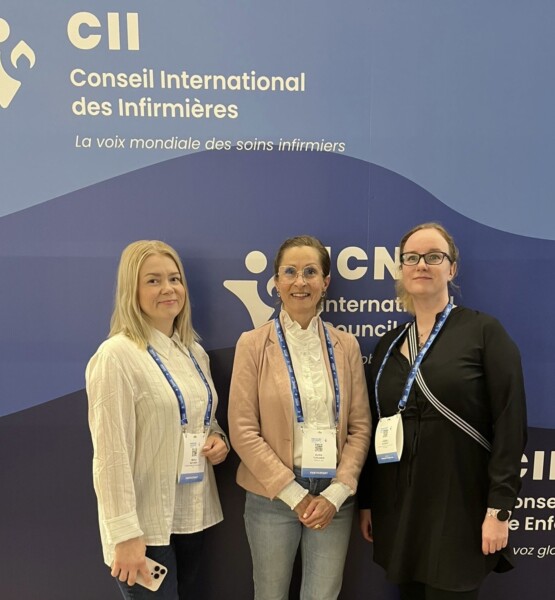 Kolme naista seisoo konferenssissa sinisen taustan edessä, jossa on Conseil International des Infirmièresin ja International Council of Nursesin logot. Heillä kaikilla on virkamerkit ja he katsovat kameraan hymyillen hieman.