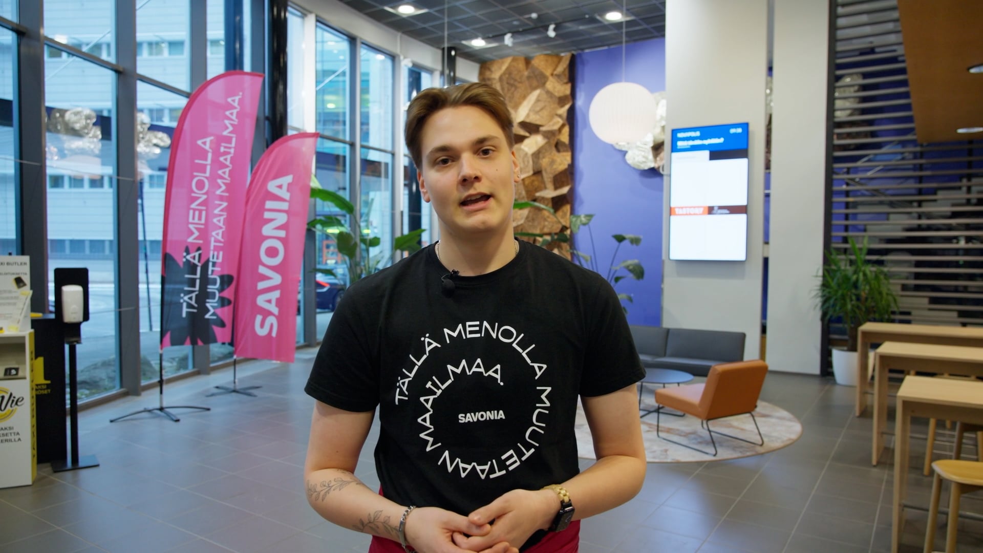 Henkilö seisoo modernissa aulassa, jossa on suuret ikkunat, vaaleanpunaiset Savonia-liput, digitaalinen näyttö ja istuimia. Henkilöllä on yllään musta Savonia-paita ja hän katsoo kameraan päin.