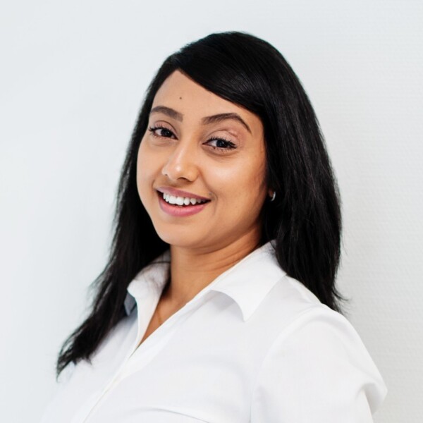 Nainen, jolla on pitkät suorat mustat hiukset ja valkoinen kauluspaita, hymyilee kameralle vaaleaa taustaa vasten.