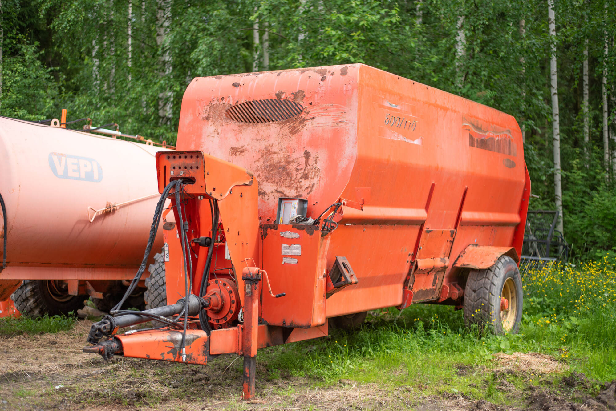 Vanha, suuri oranssi maataloudessa käytettävä sekoitin tai rehuvaunu, jossa on näkyvää likaa ja kulumaa, seisoo nurmikolla tiheiden vihreiden puiden edessä. Vasemmalla näkyy osittain toinen oranssi säiliö, jossa lukee VEPI.