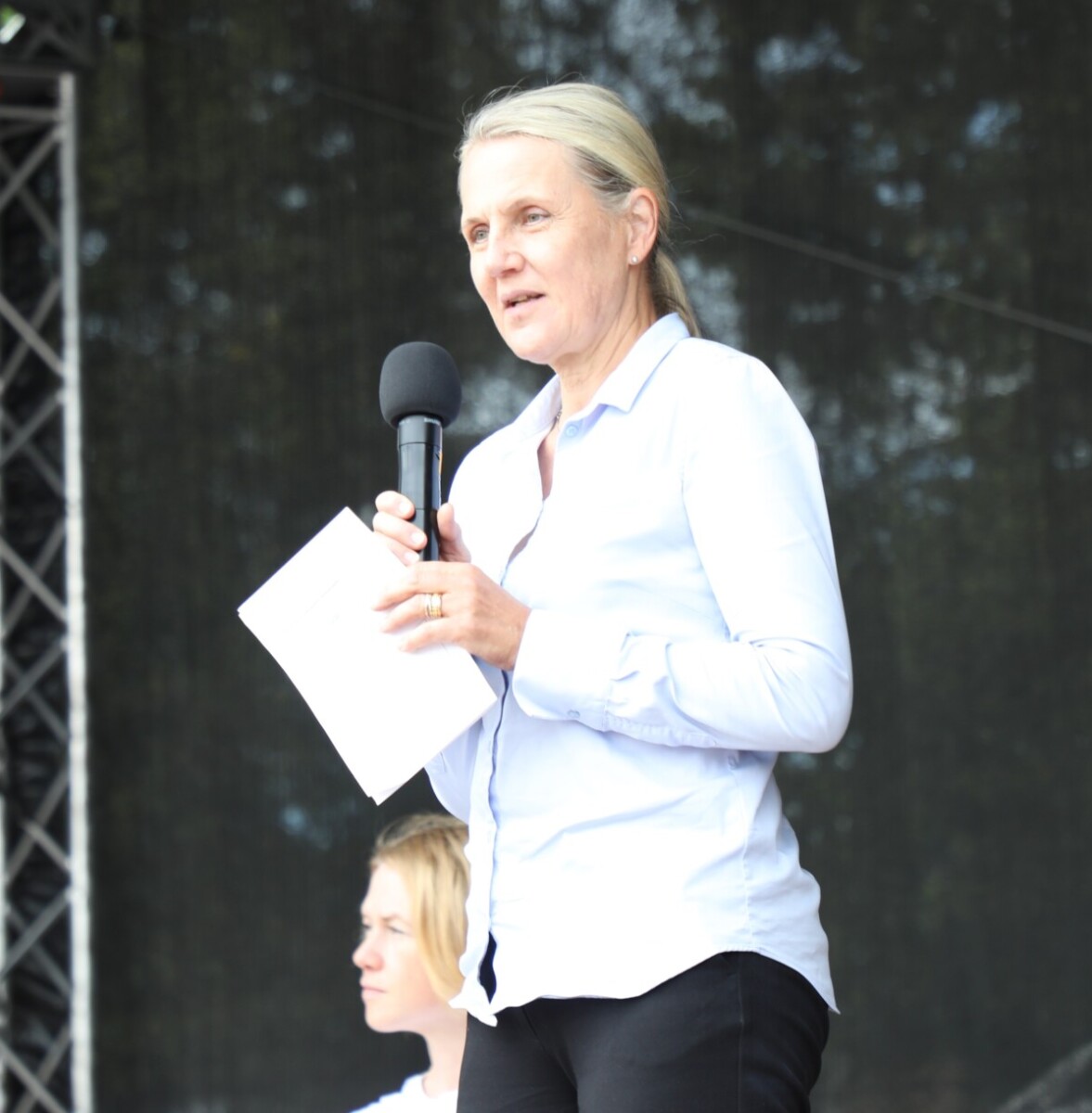 Valkoiseen puseroon pukeutunut nainen puhuu mikrofoniin ulkoilmalavalla, kädessään papereita. Hänen takanaan istuu kaksi muuta naista, ja taustalla näkyy tummia näyttämölaitteita.
