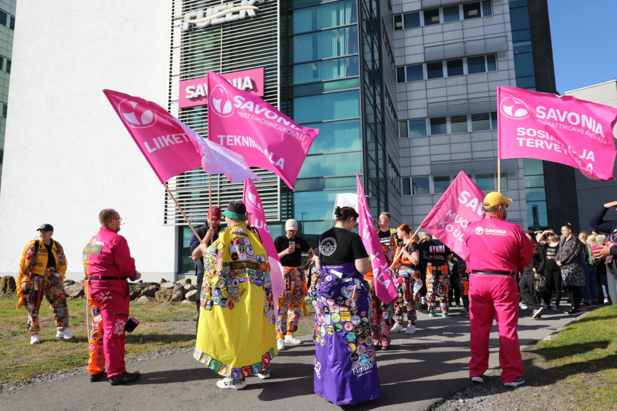 Joukko värikkäisiin haalareihin pukeutuneita ihmisiä seisoo modernin rakennuksen ulkopuolella ja heiluttaa kirkkaan vaaleanpunaisia lippuja, joissa lukee "SAVONIA". Joillakin ihmisillä on koristellut haalarit, ja kohtaus vaikuttaa juhlavalta ja vilkkaalta.