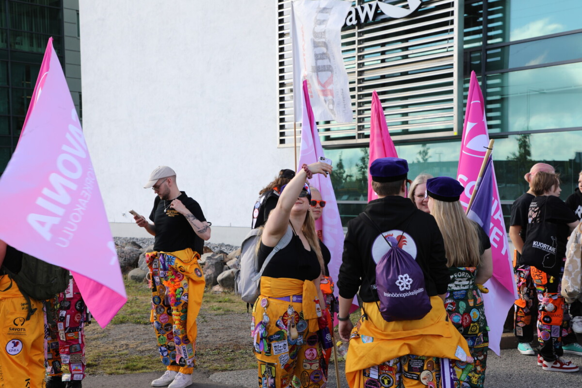 Joukko värikkäisiin, laastaripintaisiin housuihin ja hattuihin pukeutuneita ihmisiä pitelee suuria vaaleanpunaisia ja valkoisia lippuja modernin rakennuksen ulkopuolella ja kokoontuu ilmeisesti opiskelijatapahtumaan.