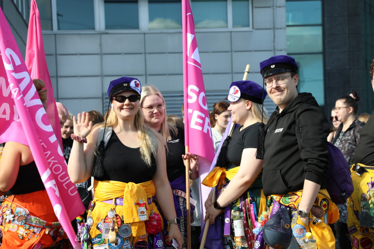 Joukko hymyileviä ihmisiä värikkäissä asuissa ja hatuissa pitelee vaaleanpunaisia lippuja ulkoilmatapahtumassa. Yksi henkilö vilkuttaa kameralle. Taustalla näkyy moderni rakennus.