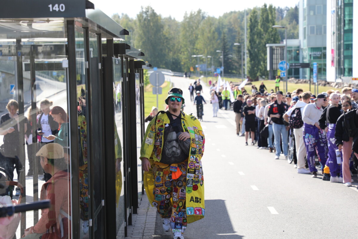Henkilö, jolla on kirkkaan keltainen takki, joka on peitetty värikkäillä laikuilla, kävelee bussipysäkin ohi, kun pitkä jono ihmisiä, joista monilla on myös koristellut haalarit, seisoo aurinkoisella kaupunkikadulla. Taustalla näkyy puita ja moderneja rakennuksia.