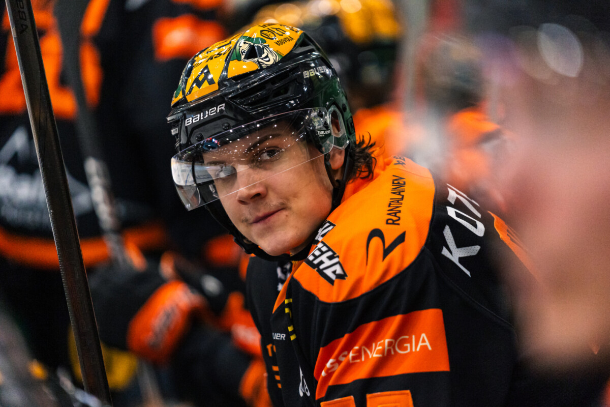 Jääkiekkoilija, jolla on musta-oranssi peliasu ja kypärä, istuu penkillä ja katsoo kohti kameraa. Taustalla näkyy muita samanlaisiin peliasuihin pukeutuneita pelaajia.