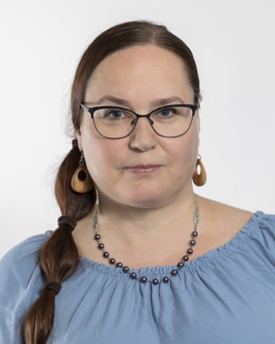 Nainen, jolla on pitkät ruskeat hiukset sivupunoksessa, silmälasit, puiset korvakorut, helmikaulakoru ja vaaleansininen olkatop, poseeraa valkoista taustaa vasten.