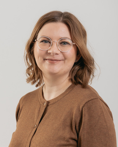 Nainen, jolla on hartioiden pituiset vaaleanruskeat hiukset, pyöreät silmälasit ja ruskea napillinen toppi, hymyilee lempeästi kameralle vaaleaa taustaa vasten.