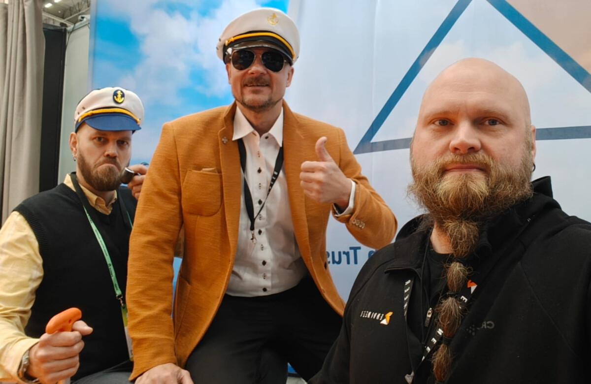 Kolme miestä poseeraa sisätiloissa, kahdella on valkoiset kapteeninhatut ja yhdellä aurinkolasit ja oranssi takki, ja he osoittavat peukkua ylöspäin. Taustalla on taivas ja geometrinen kuvio.