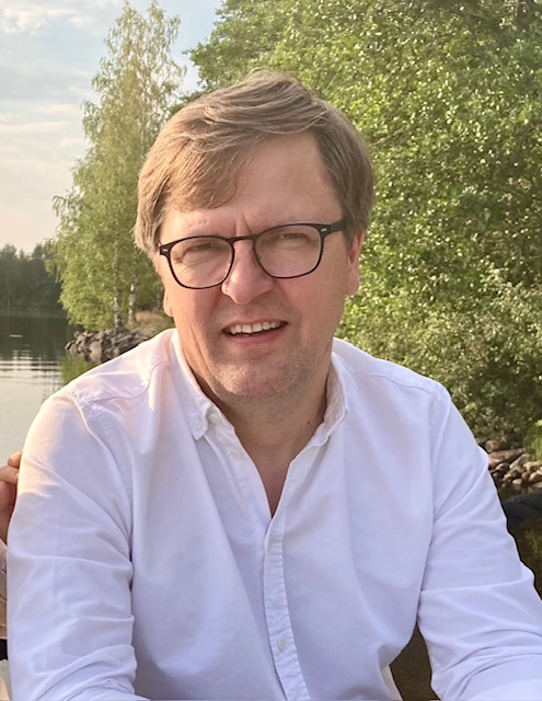 Silmälasipäinen ja valkoiseen paitaan pukeutunut mies istuu ulkona rauhallisen järven rannalla, vehreiden puiden ympäröimänä ja osittain pilvisen taivaan alla.