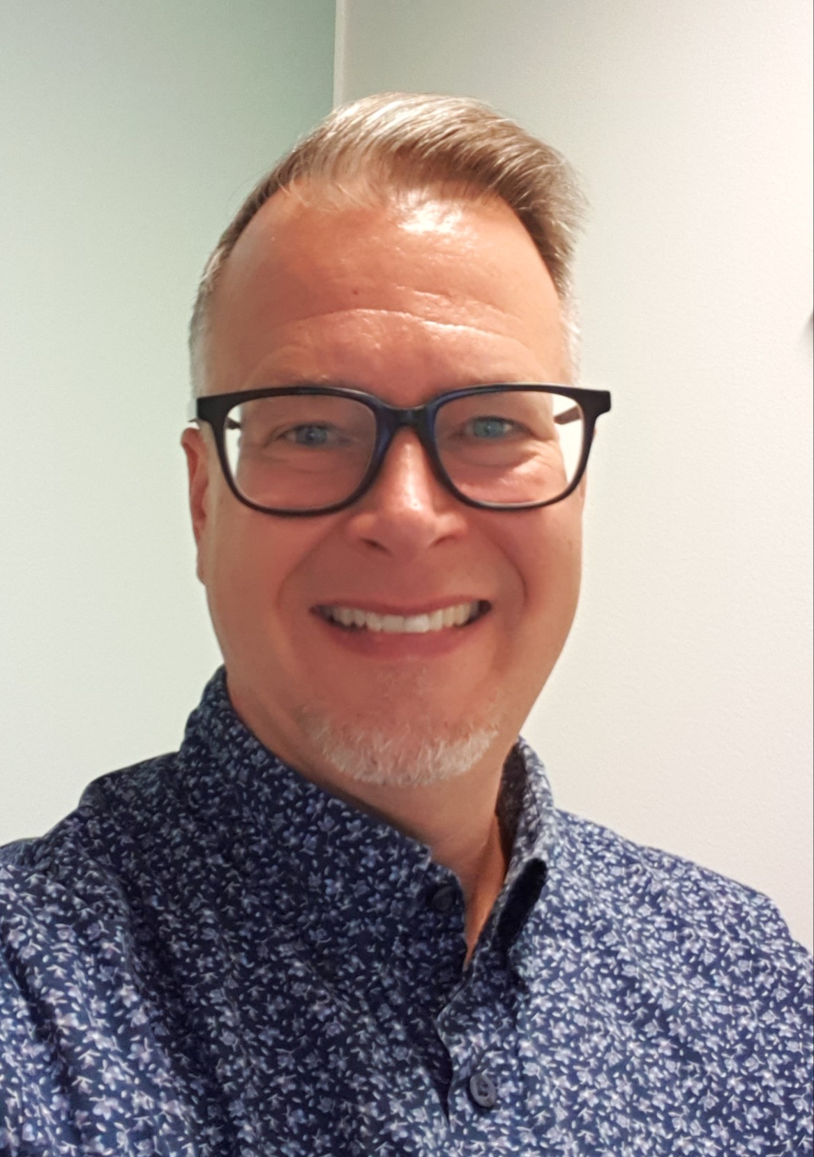 Hymyilevä mies, jolla on lyhyet vaaleat hiukset, silmälasit ja lyhyt parta, ja jolla on sininen kuvioitu paita, seisoo vaaleaa seinää vasten.