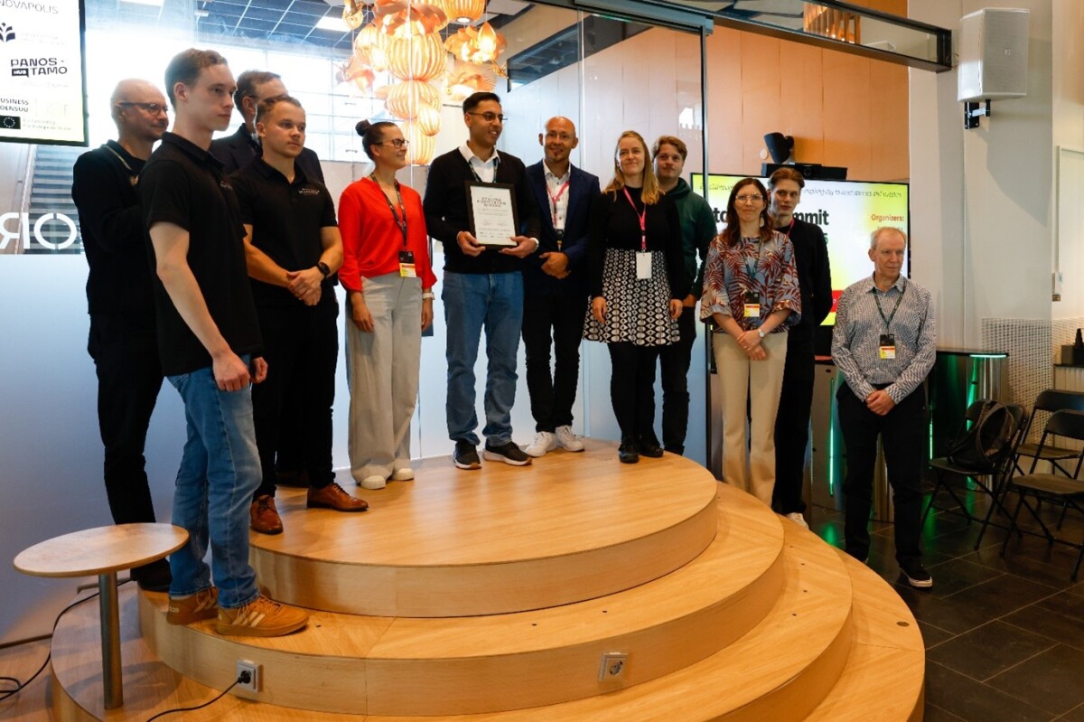 Joukko ihmisiä poseeraa puisella näyttämöllä, ja yksi henkilö on keskellä pitelemässä muistolaattaa. Jotkut henkilöt hymyilevät, ja muutamilla on kaulanauhat. Taustalla näkyy moderni valaistus ja lasiseinä.