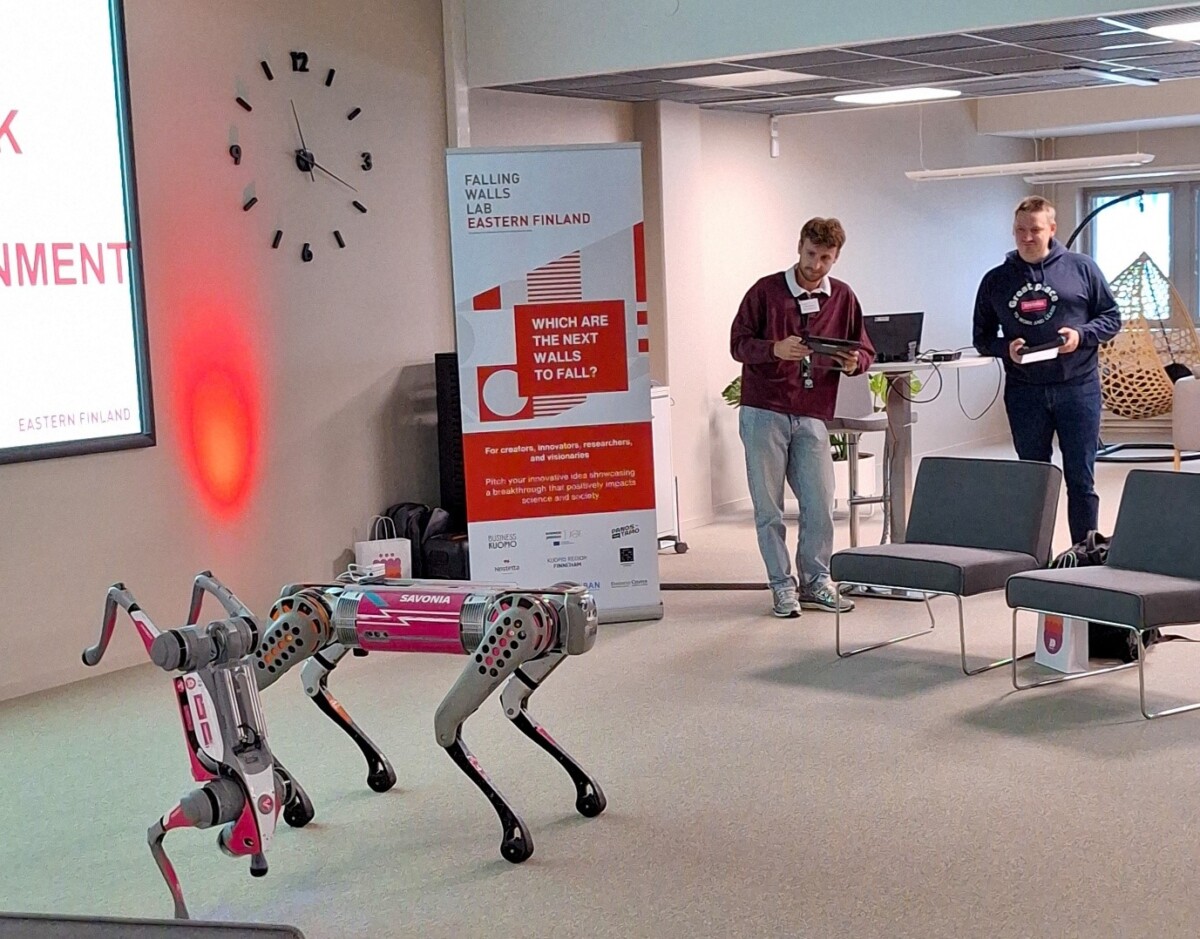 Kaksi ihmistä käyttää nelijalkaista robottikoiraa modernissa huoneessa. Heidän takanaan on banneri, jossa on tekstiä ja suuri seinäkello. Näyttää siltä, että kyseessä on tekninen esittely tai tapahtuma.