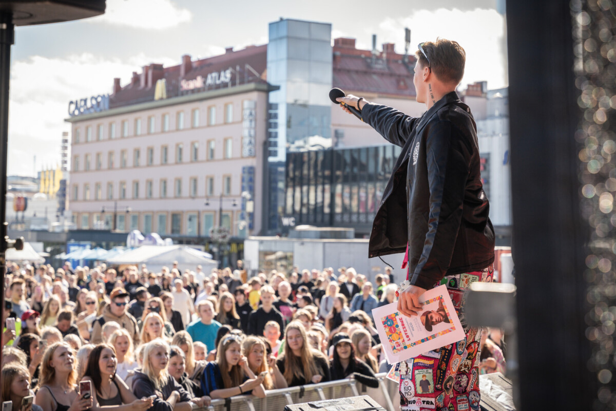 Esiintyjä, jolla on kädessään mikrofoni ja värikäs kansio, puhuu suurelle, hymyilevälle yleisölle ulkoilmatapahtumassa kaupungin aukiolla, taustalla näkyy rakennuksia ja telttoja.