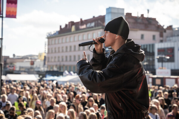 Henkilö, jolla on musta pipo ja nahkatakki, esiintyy lavalla mikrofonin kanssa ja on suurelle ulkoilmayleisölle päin kaupungin aukiolla päivällä.