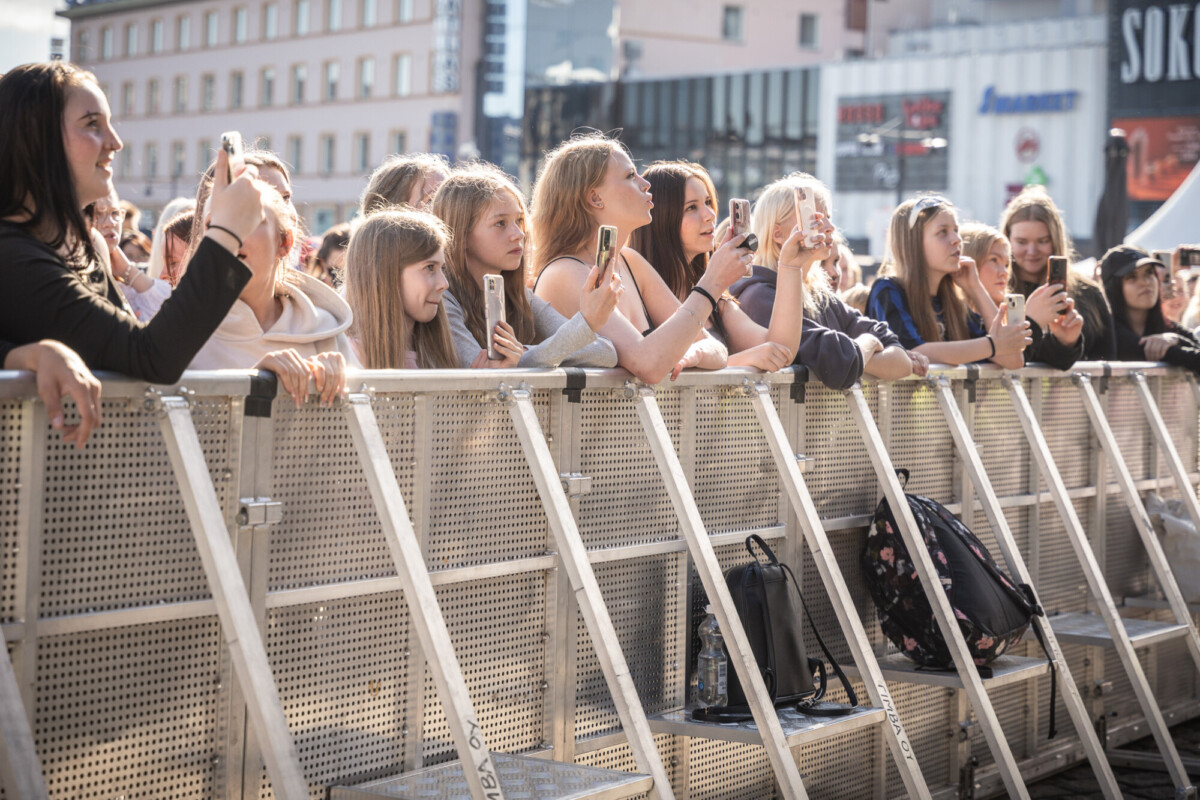 Joukko innostuneita nuoria seisoo metalliesteen takana ulkoilmatapahtumassa, ja monet pitävät älypuhelimia kädessään ottaakseen valokuvia tai videoita. Kohtaus sijoittuu kaupunkiin, jonka taustalla on rakennuksia.