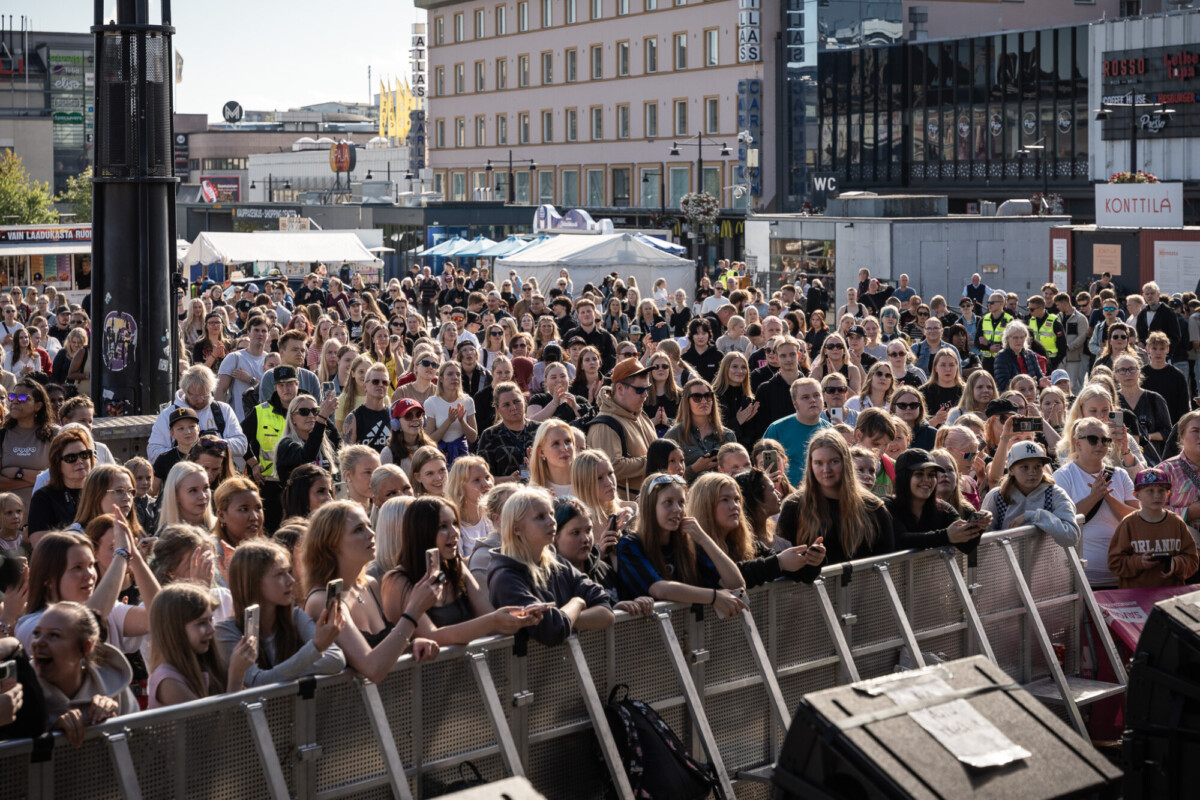 Suuri ihmisjoukko seisoo tiiviisti yhdessä ulkoilmatapahtumassa kaupungin aukiolla lavaa vasten. Kohtaus on valoisa ja aurinkoinen, ja taustalla näkyy rakennuksia ja markkinateltta.