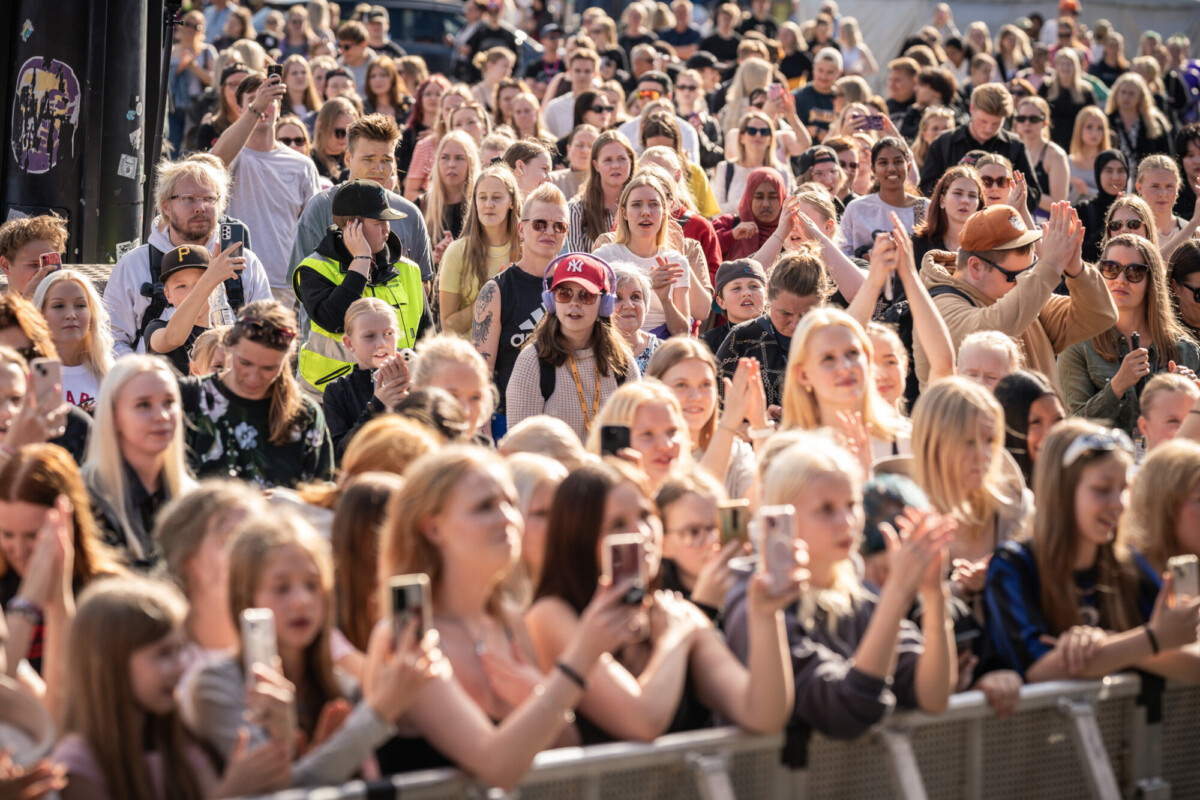 Suuri ihmisjoukko, monet pitävät puhelimia kädessään ja taputtavat, kokoontuu ulkona aurinkoisessa tapahtumassa tai konsertissa. Yleisö vaikuttaa sitoutuneelta ja innostuneelta, ja siinä näkyy eri-ikäisiä ja eri ilmeisiä ihmisiä.