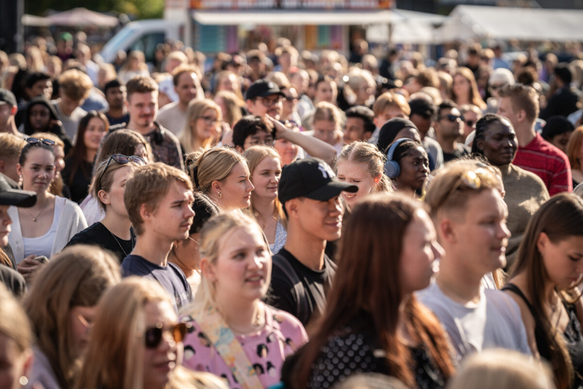 Suuri, erilainen ihmisjoukko kokoontuu ulkona päivänvalossa, jotkut hymyilevät ja puhuvat, toiset katsovat eteenpäin. Taustalla näkyy kojuja ja telttoja, jotka viittaavat festivaaliin tai julkiseen tapahtumaan.