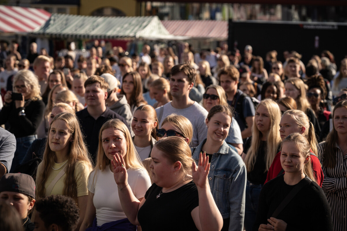 Suuri joukko ihmisiä, enimmäkseen nuoria aikuisia, seisoo ulkona auringonvalossa. He näyttävät katselevan tapahtumaa, ja jotkut hymyilevät ja toiset näyttävät tarkkaavaisilta. Taustalla näkyy markkinakojuja ja rakennuksia.