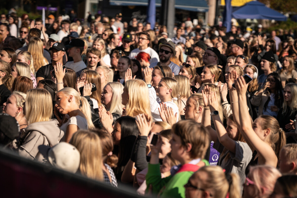 Suuri ihmisjoukko ulkona päivänvalossa, monet nostavat kädet pystyyn ja nauttivat tapahtumasta tai konsertista. Useimmat katsovat samaan suuntaan, ja ilmapiiri vaikuttaa vilkkaalta ja energiseltä.