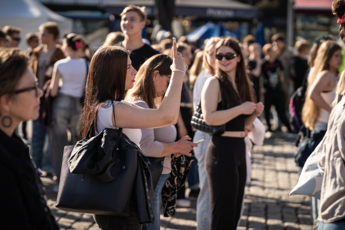 Ryhmä nuoria seisoo ulkona auringonvalossa. Yksi etualalla oleva nainen pitää puhelinta kädessään ja näyttää ottavan valokuvaa tai videota. Muut hymyilevät ja keskustelevat, ja jotkut katsovat puhelimiinsa. Alue on vilkas ja vilkas.