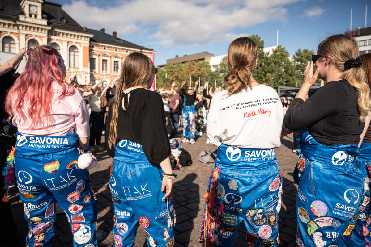 Joukko sinisiin haalareihin ja värikkäisiin laastareihin pukeutuneita opiskelijoita seisoo kaupungin aukiolla ja katsoo väkijoukkoon kädet kohotettuna. Kohtaus on vilkas ja ulkona, ja taustalla on historiallisia rakennuksia ja puita.