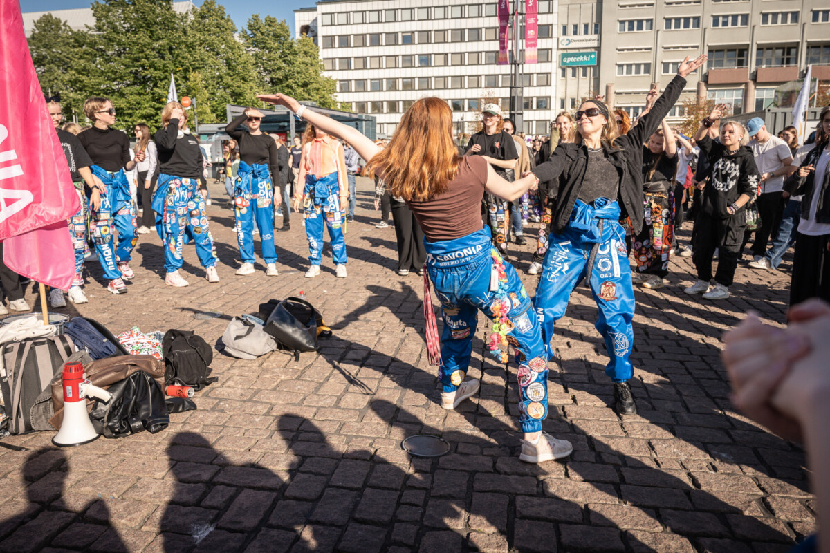Joukko sinisiin haalareihin pukeutuneita opiskelijoita tanssii ja juhlii ulkona kaupungin aukiolla muiden ihmisten ja rakennusten ympäröimänä aurinkoisena päivänä.