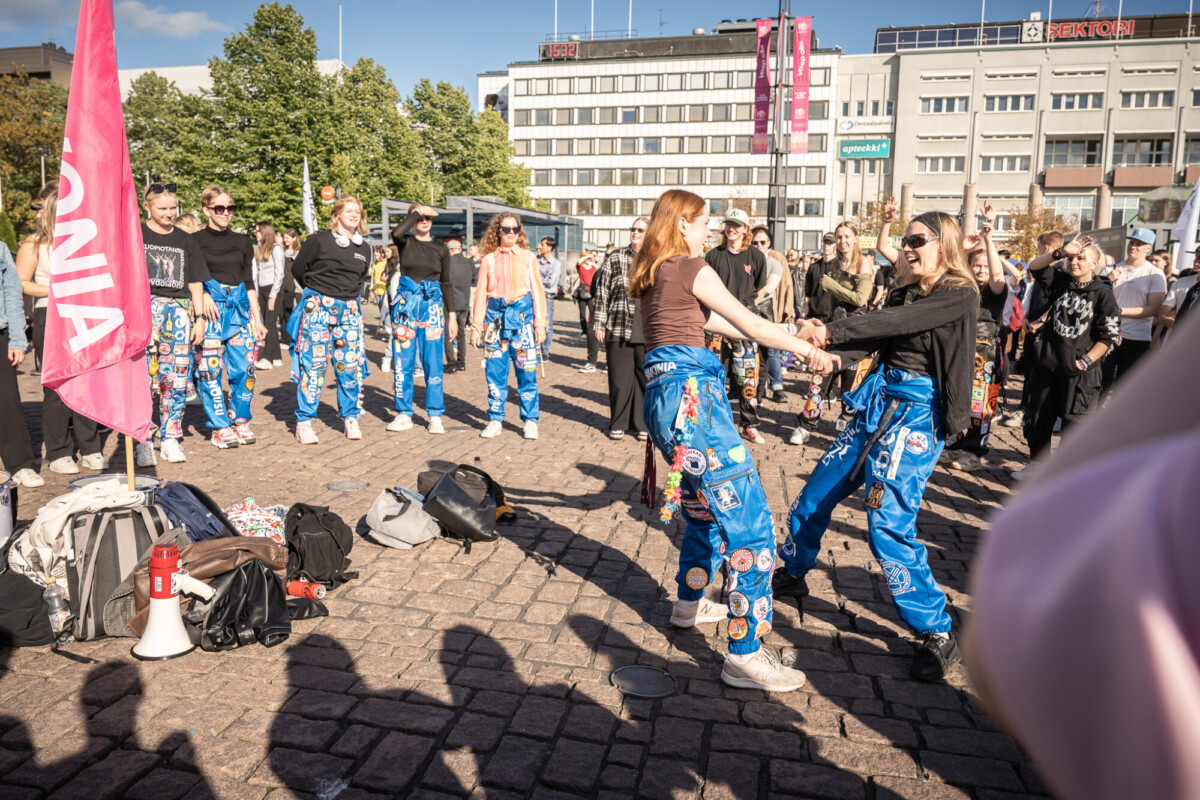 Joukko värikkäisiin haalareihin pukeutuneita nuoria seisoo kaupungin aukiolla. Kaksi naista tanssii keskellä, kun muut katselevat ja hymyilevät. Laukut ja megafoni ovat maassa. Taustalla kirkas, aurinkoinen sää ja rakennuksia.