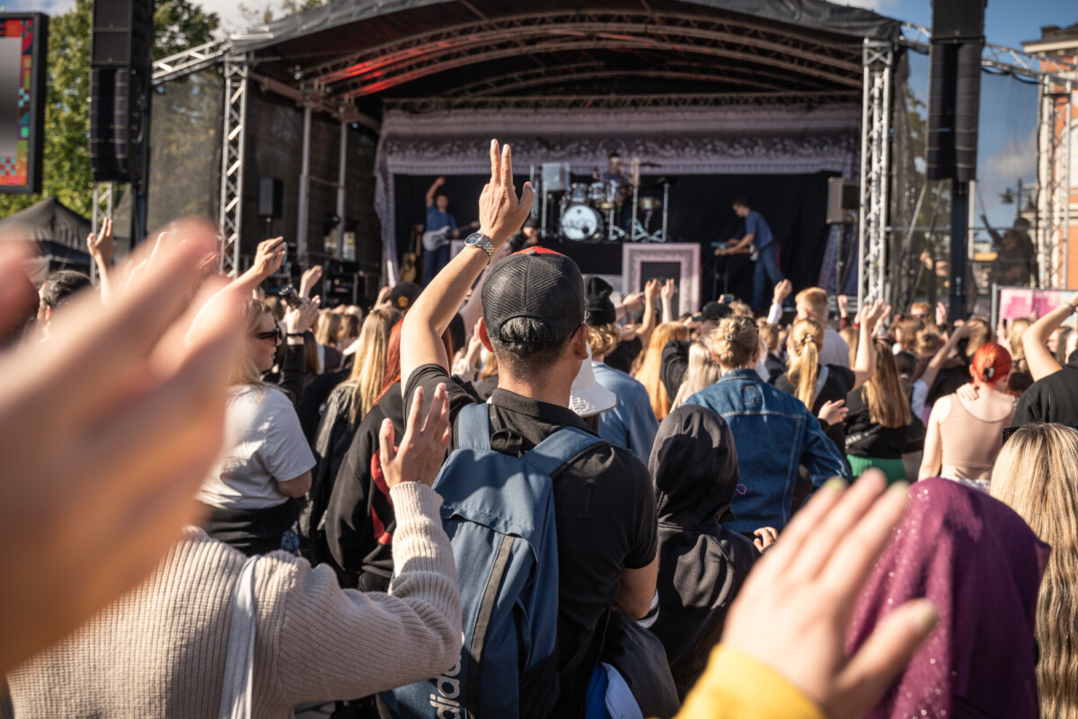 Ihmisjoukko ulkoilmalavalla konsertissa, jotkut nostavat kätensä ylös. Muusikot esiintyvät lavalla katoksen alla, ja yleisö nauttii aurinkoisesta tapahtumasta.