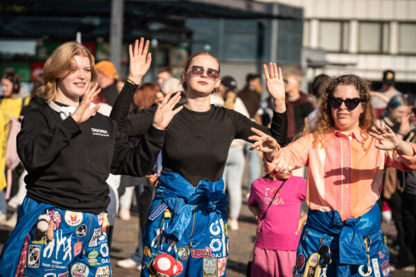 Kolme nuorta naista seisoo ulkona väkijoukossa ja nostaa kätensä ilmaan. Heillä on yllään kirkkaan siniset haalarit, joita koristavat erilaiset laastarit ja merkit. Auringonvalo paistaa heihin, ja taustalla näkyy ihmisiä.