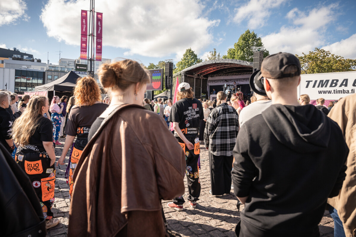 Yleisö kokoontuu ulkona tapahtuman lavalle. Monilla ihmisillä on yllään värikkäät haalarit, joissa on laastareita. Taivas on osittain pilvinen, ja taustalla näkyy puita ja rakennuksia.