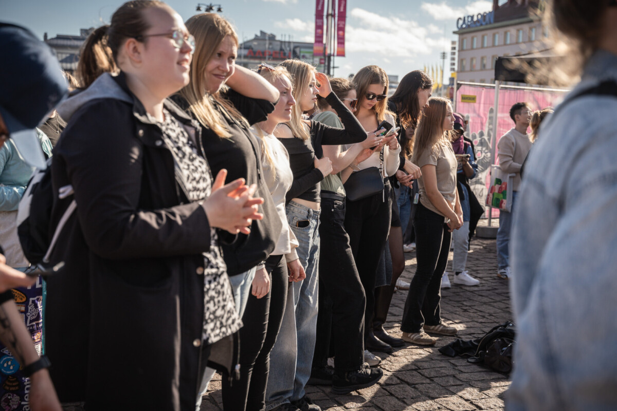 Joukko ihmisiä seisoo ulkona aurinkoisella kaupunkialueella, jotkut taputtavat, toiset ottavat valokuvia tai katselevat puhelimiaan. Taustalla näkyy rakennuksia ja banderolleja.