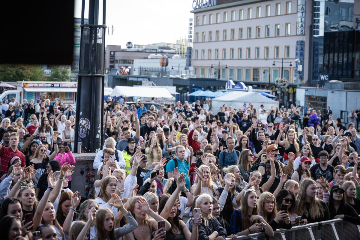 Suuri ihmisjoukko ulkona konsertissa tai tapahtumassa, monilla kädet kohotettuna, seisoo lavan edessä, ja taustalla näkyy rakennuksia, telttoja ja kylttejä.