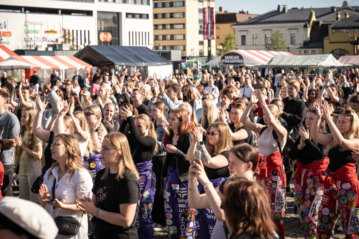 Suuri joukko nuoria ihmisiä, joista monilla on värikkäät haalarit ja laastarit, seisoo ulkona festivaaleilla tai tapahtumissa taputtaen ja hurraa-huutoja torikojujen ja kaupungin rakennusten edessä aurinkoisella säällä.