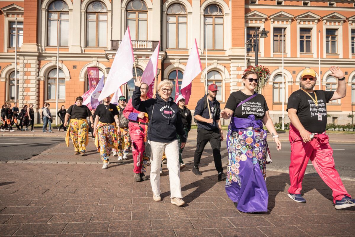 Joukko värikkäisiin vaatteisiin ja koristeltuihin haalareihin pukeutuneita ihmisiä kävelee paraatissa, jotkut vilkuttavat ja hymyilevät, taustalla historiallisia rakennuksia ja lippuja.