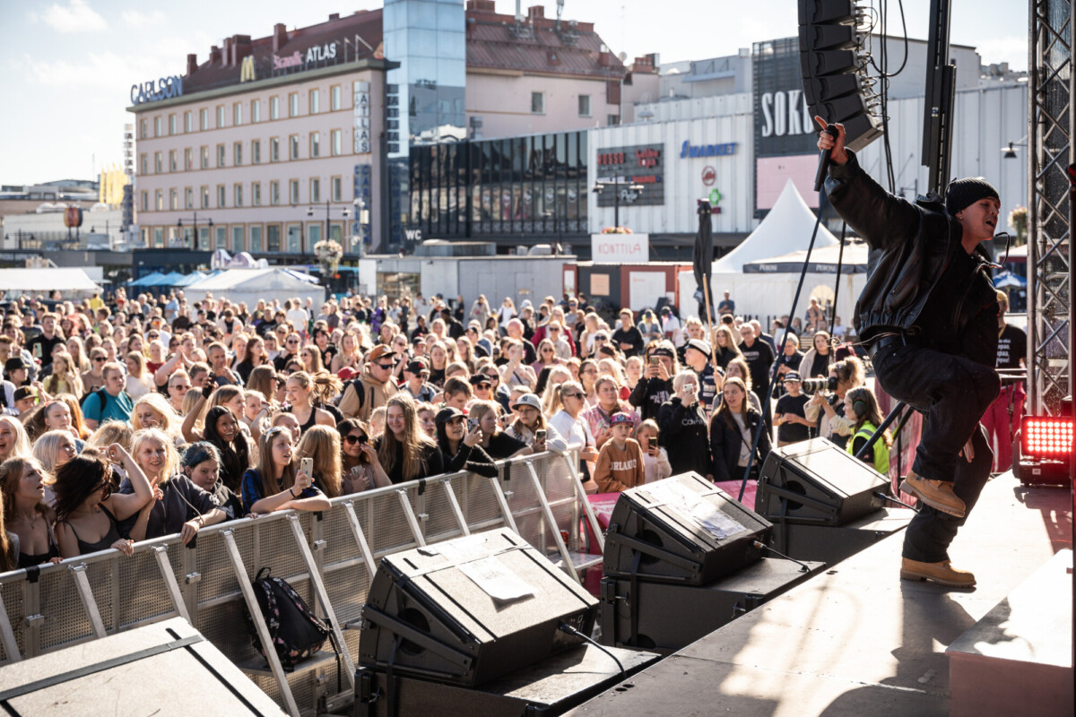 Esiintyjä lavalla virittää suurta ulkoilmayleisöä musiikkifestivaaleilla. Yleisö on sitoutunut, monet nostavat kätensä ylös, ja taustalla näkyy kaupungin rakennuksia aurinkoisella taivaalla.