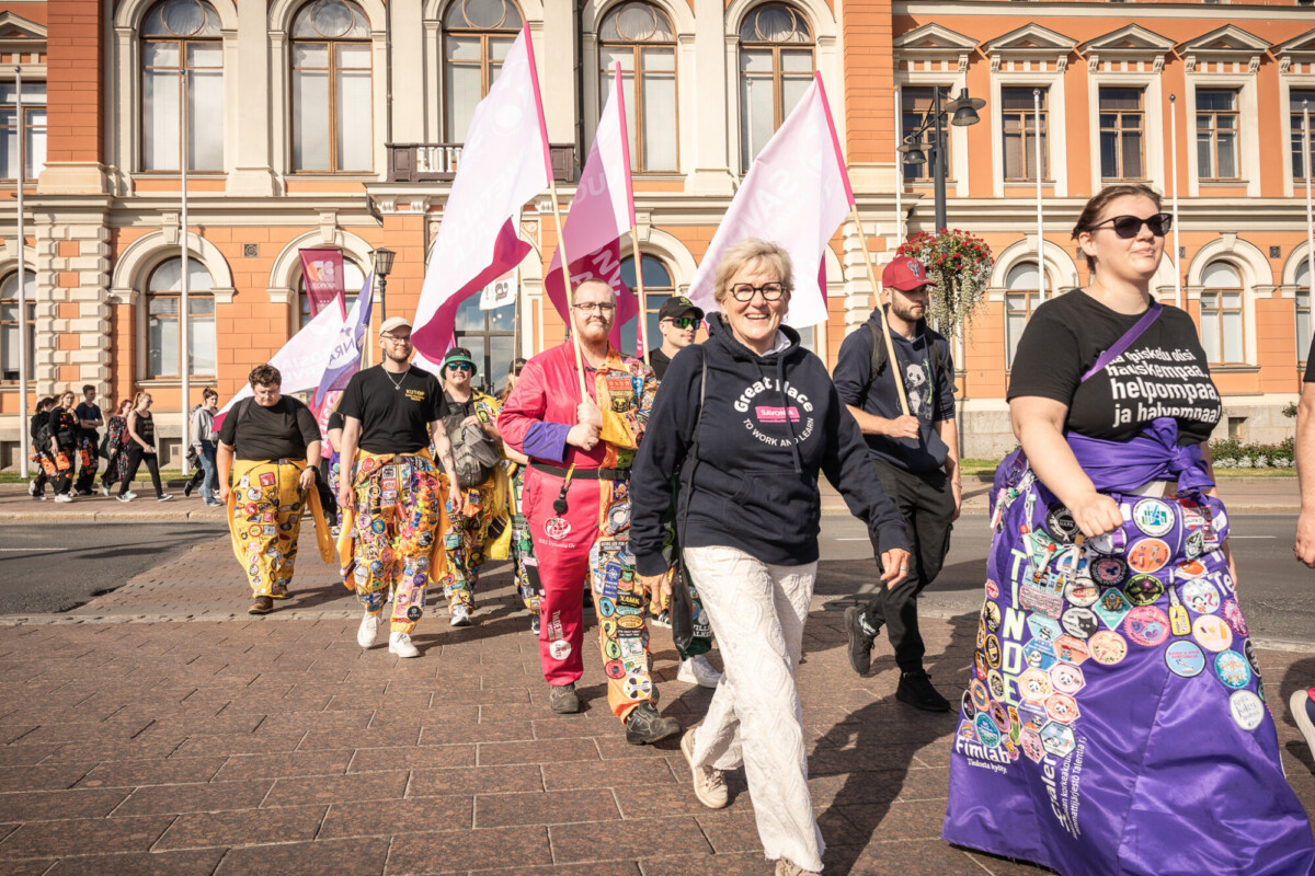 Joukko ihmisiä marssii ulkona värikkäisiin haalareihin pukeutuneena, laastareilla ja nastoilla koristeltuina, lippuja kantaen ja hymyillen. He ovat koristeellisen rakennuksen edessä, jossa on suuret ikkunat ja pylväät.