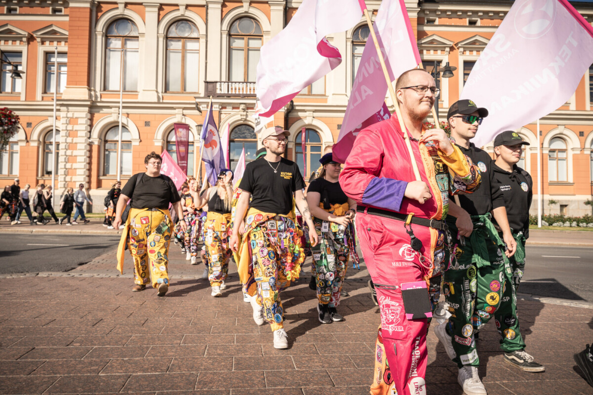 Joukko värikkäisiin haalareihin pukeutuneita ihmisiä, joilla on laastareita ja pinssejä, marssivat paraatissa, joillakin on lippuja kädessään, ja taustalla näkyy historiallinen rakennus.