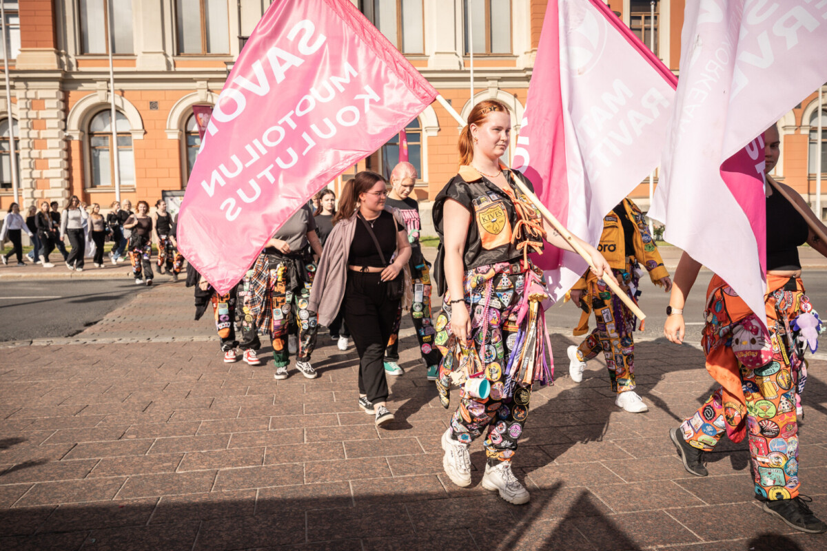 Ryhmä värikkäisiin haalareihin pukeutuneita ja laastareilla koristeltuja ihmisiä marssii ulkona, kädessään suuret vaaleanpunaiset liput, joissa on teksti. He ovat historiallisen oranssin ja kermanvärisen rakennuksen edessä aurinkoisena päivänä.