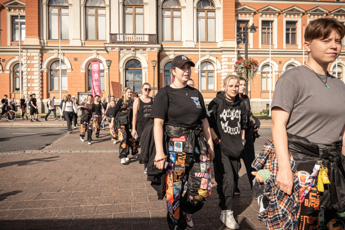 Joukko nuoria, joilla on koristellut housut ja rennot vaatteet, kävelee yhdessä kaupungin kadulla historiallisen rakennuksen edessä, jossa on suuret ikkunat ja oranssit seinät.