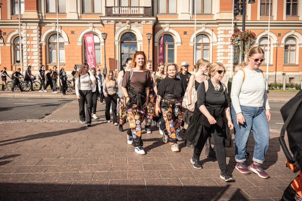 Joukko nuoria, joista monilla on tummat housut, joissa on värikkäitä laikkuja, kävelee yhdessä aurinkoisella kadulla suuren, koristeellisen rakennuksen edessä, jonka seinät ovat oranssin ja beigen väriset.
