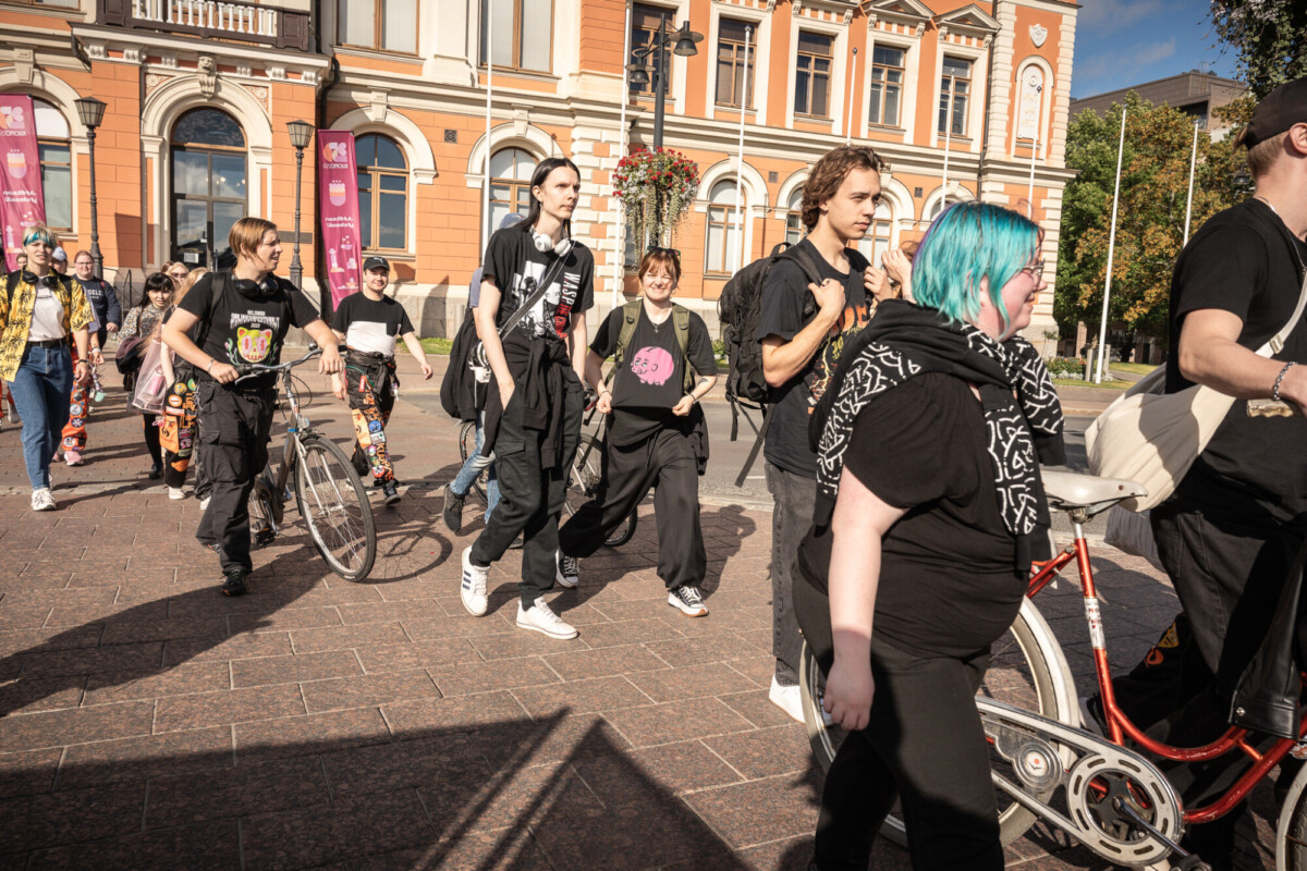 Joukko nuoria, joilla on värikkäät hiukset ja mustat vaatteet, kävelee ja pyöräilee aurinkoisella kaupunkiaukiolla, jonka taustalla on historiallisia rakennuksia.