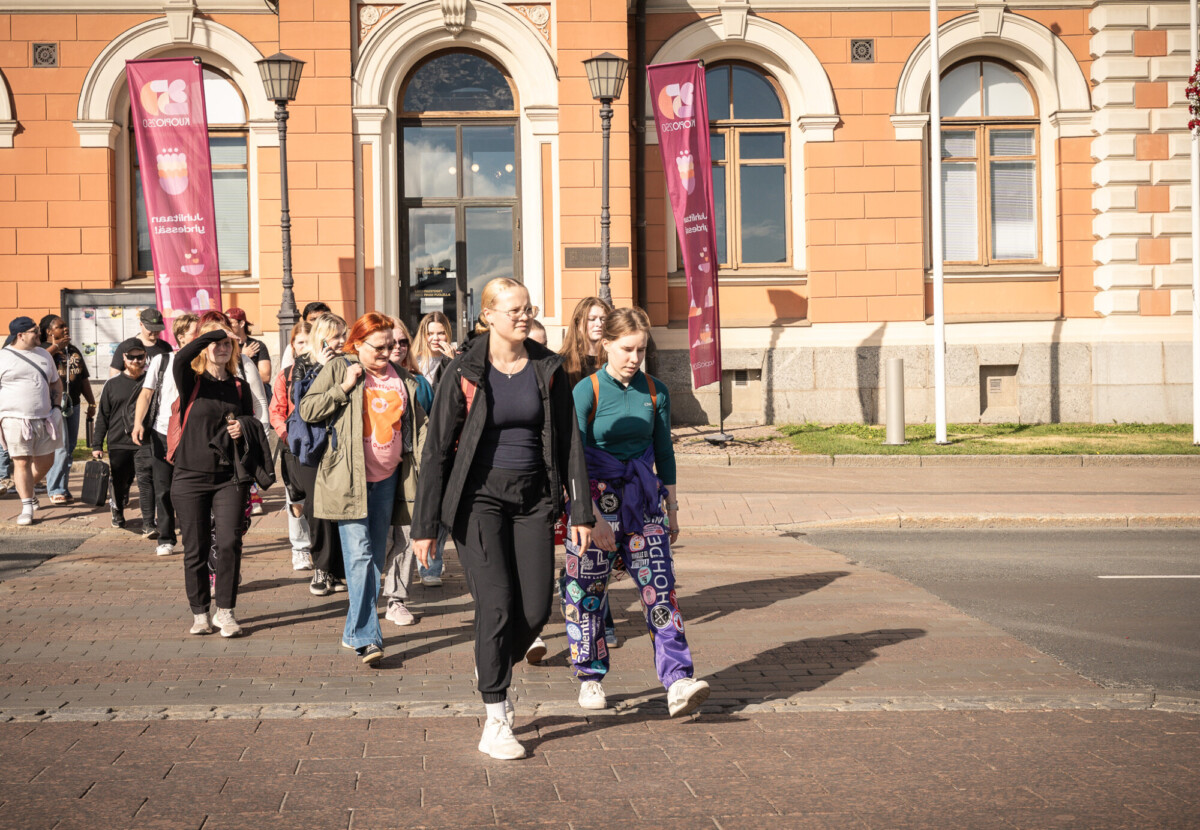 Joukko ihmisiä, enimmäkseen nuoria naisia, kävelee yhdessä aurinkoisena päivänä katua pitkin lähellä suurta oranssia rakennusta, jossa on kaarevat ikkunat ja jonka ulkopuolella roikkuu vaaleanpunaisia julisteita.